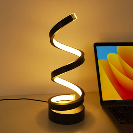 Candeeiro LED de mesa com design espiral moderno em preto, emitindo luz quente e posicionado ao lado de um portátil numa secretária.